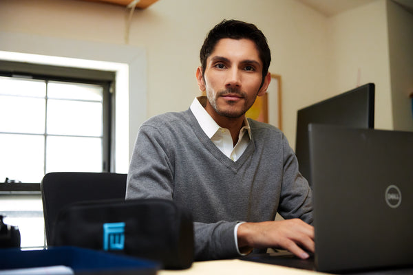 Man dressed in business casual clothing, grey sweater and white collared shirt, with dark hair and a goatee sitting in front of his laptop in a home office setting. A Fisher Wallace carrying case rests on the table near him.