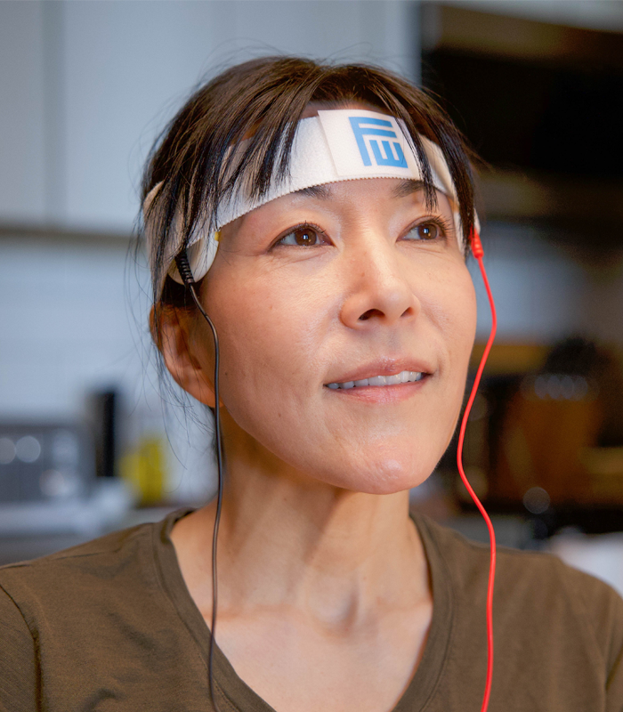 A woman with short bangs and hair tied in a ponytail is using the Fisher Wallace Stimulator. She has a smile on her face as she feels the treatment. She is wearing a vee neck olive t-shirt.