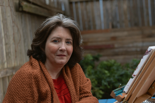 Image of an older woman with grey and brown hair sitting in her backyard, doing some amateur painting. She has on an orange sweater and red shirt.
