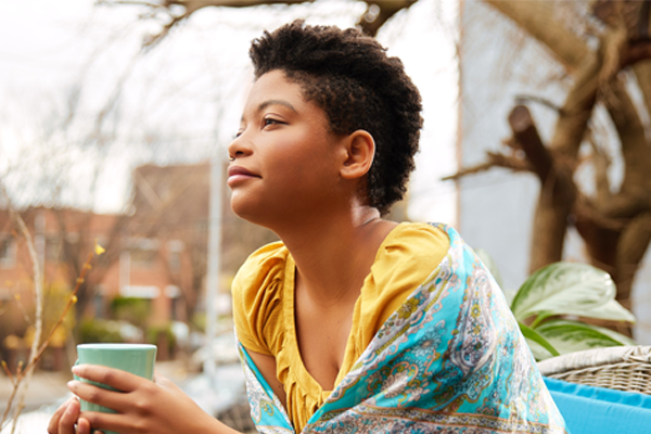 Young black woman sits outside holding a mint-colored mug. She's wearing a mustard colored shirt with a blue and white paisley shawl. Apartment buildings and a tree are in the background.