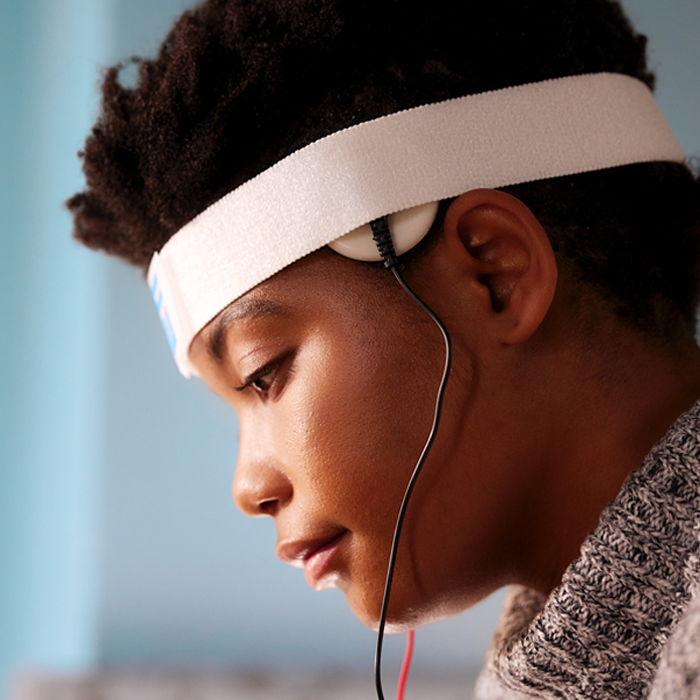 Profile view of a young black woman wearing a black and white turtleneck sweater in treatment with the Fisher Wallace stimulator. She has the headband on her head and electrodes in place above the side burns.